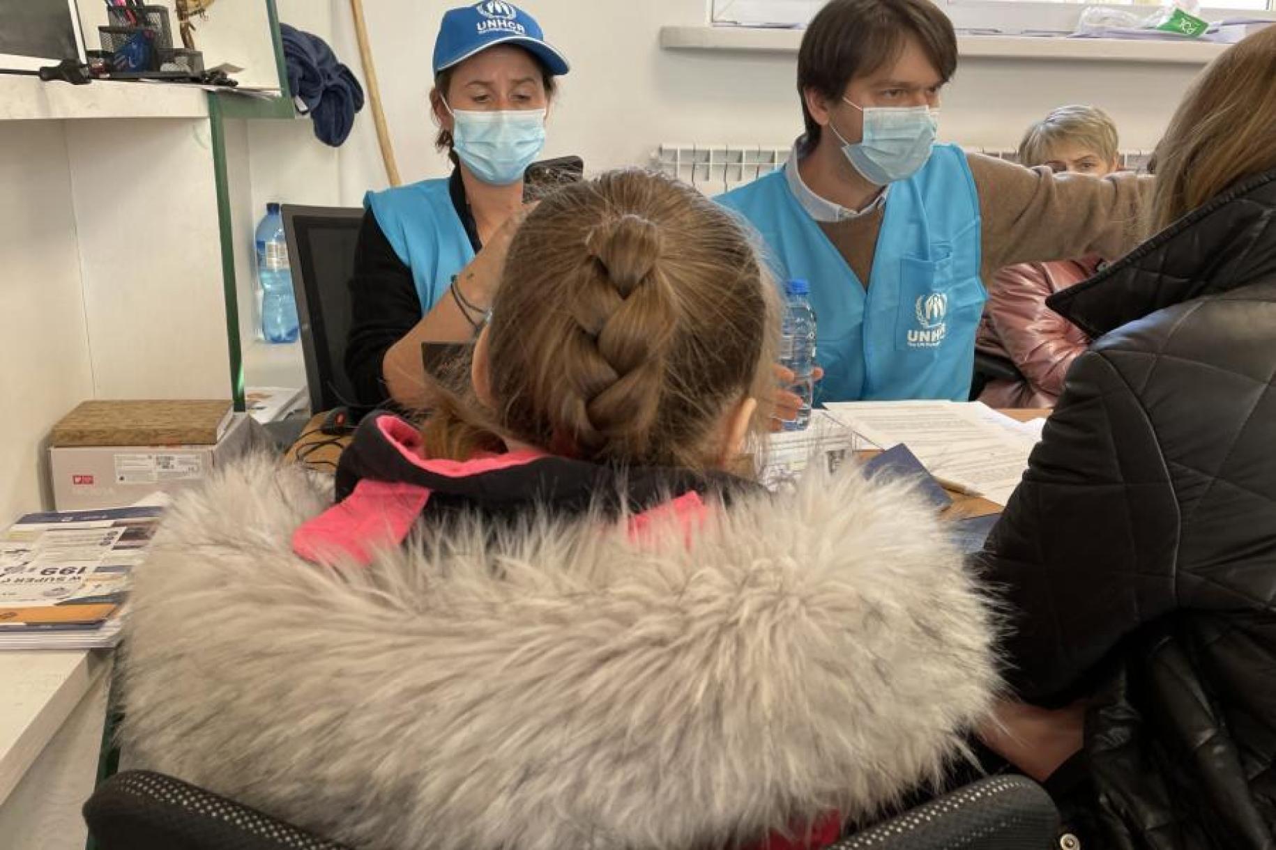 In an indoor area, two people in blue vests and wearing facemasks are having a conversation with an adult and a girl facing away from the camera and wearing warm clothes, and there is another person sitting in the back of the room.