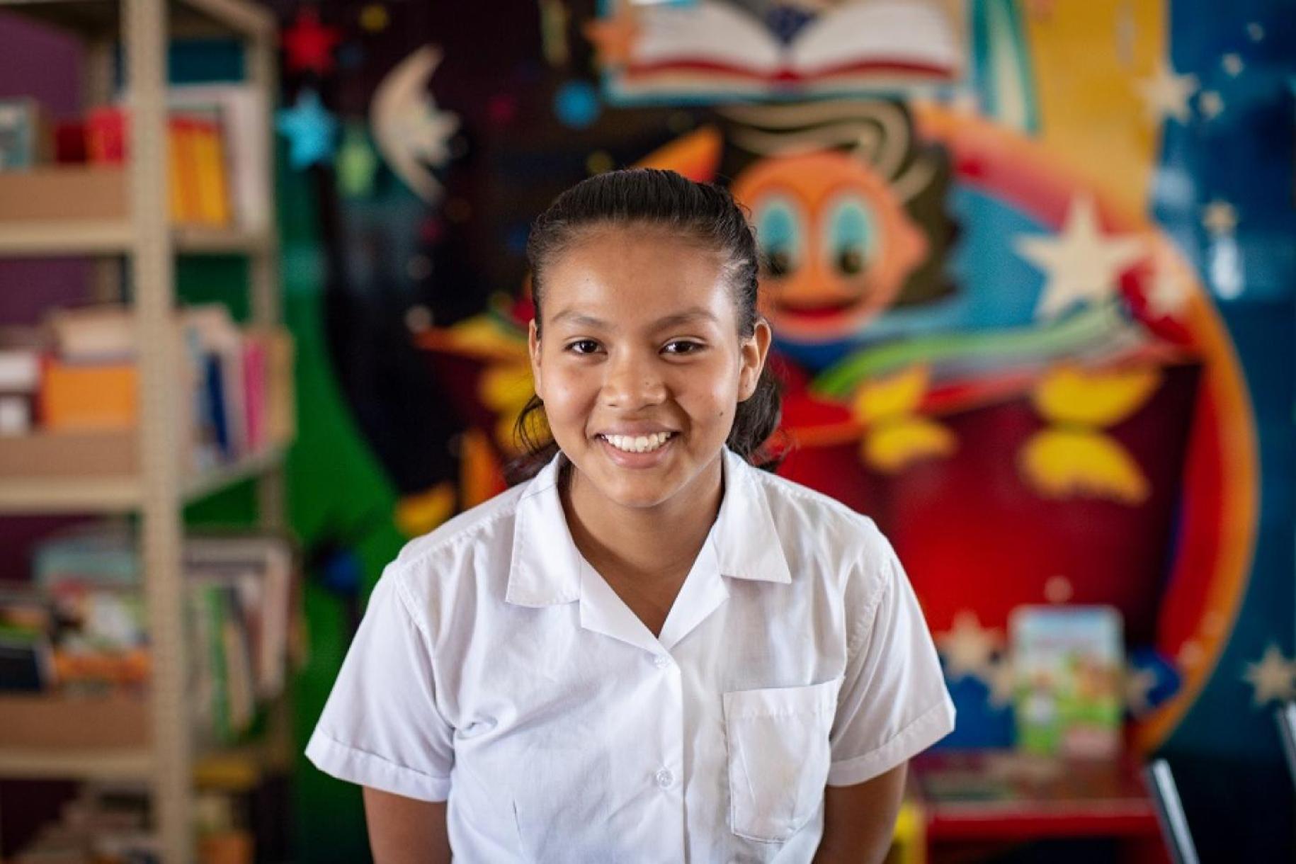 一个穿着校服的女孩对着镜头微笑，站在一幅彩色壁画和一个摆满书的书架前。