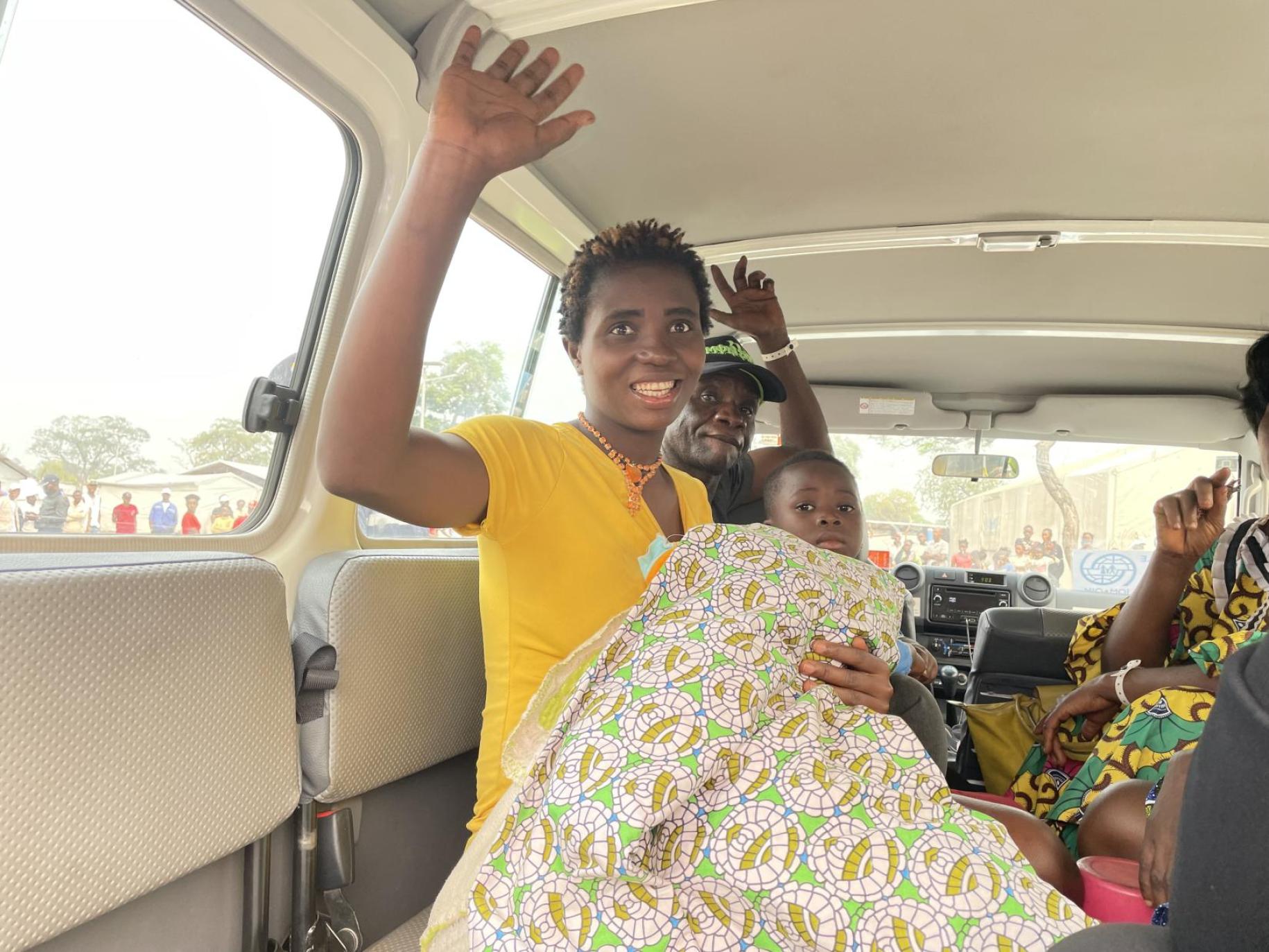 In Angola, a Congolese woman with short hair and wearing a yellow short-sleeved top sits in a vehicle with other poeple, a baby wrapped in a blanket on her knees, and waves goodbye with her right hand and smiling.