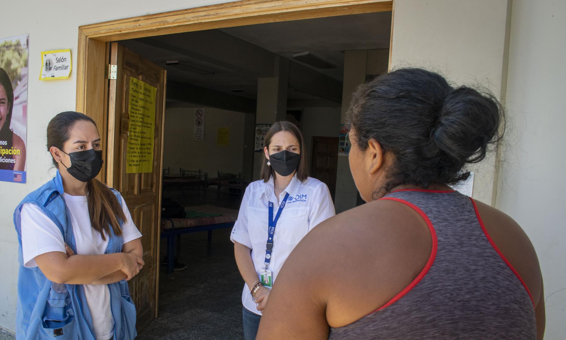 一位背对镜头站立的妇女正在与两位戴着面罩的人交谈，其中一位穿着背心，表明她是联合国系统的成员，另一位则佩戴着联合国的ID徽章。