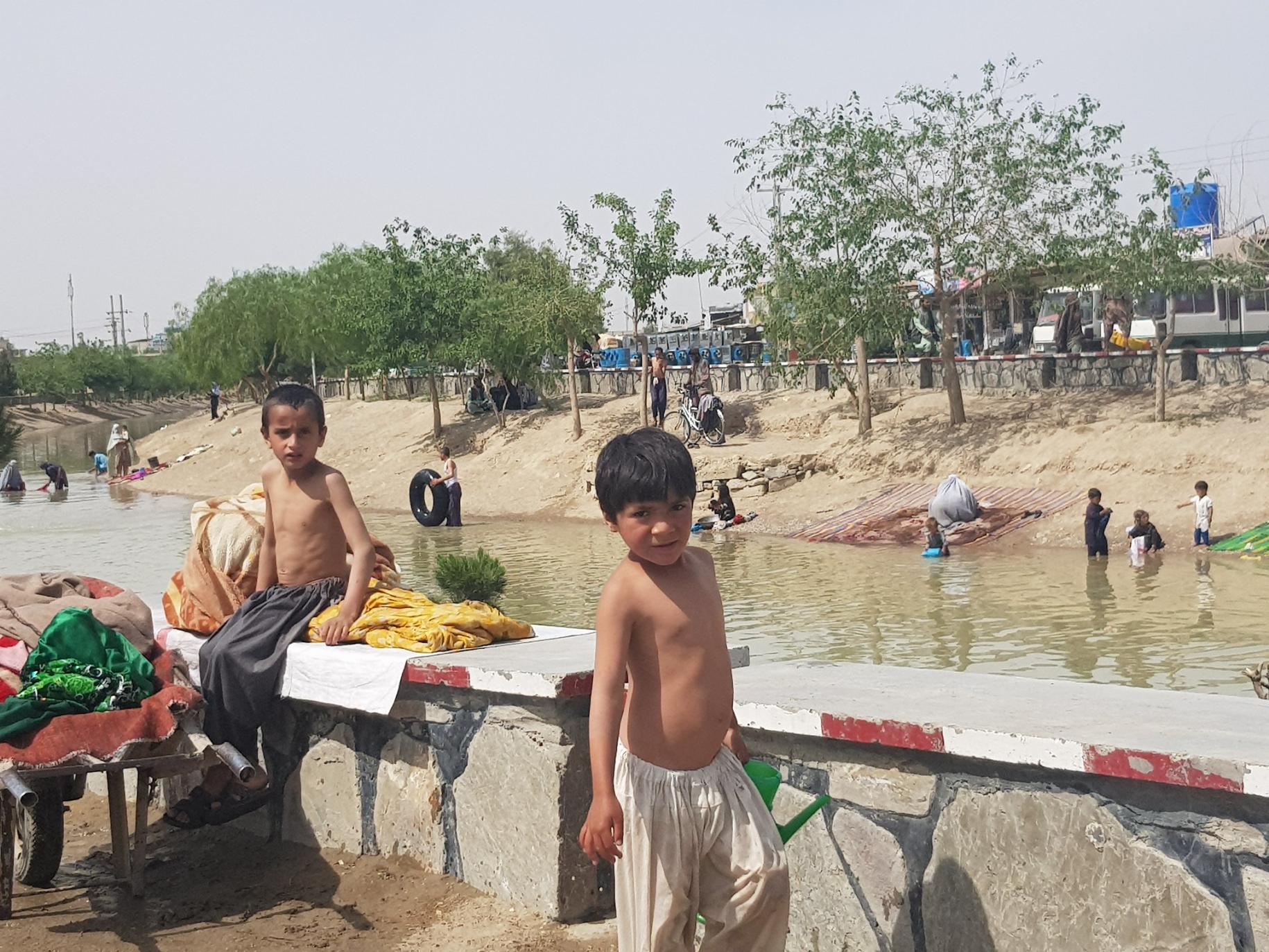 Des Afghan·e·s lavent des vêtements et des tapis dans une rivière, en famille, tandis que des enfants jouent au bord de l’eau pour se rafraîchir.