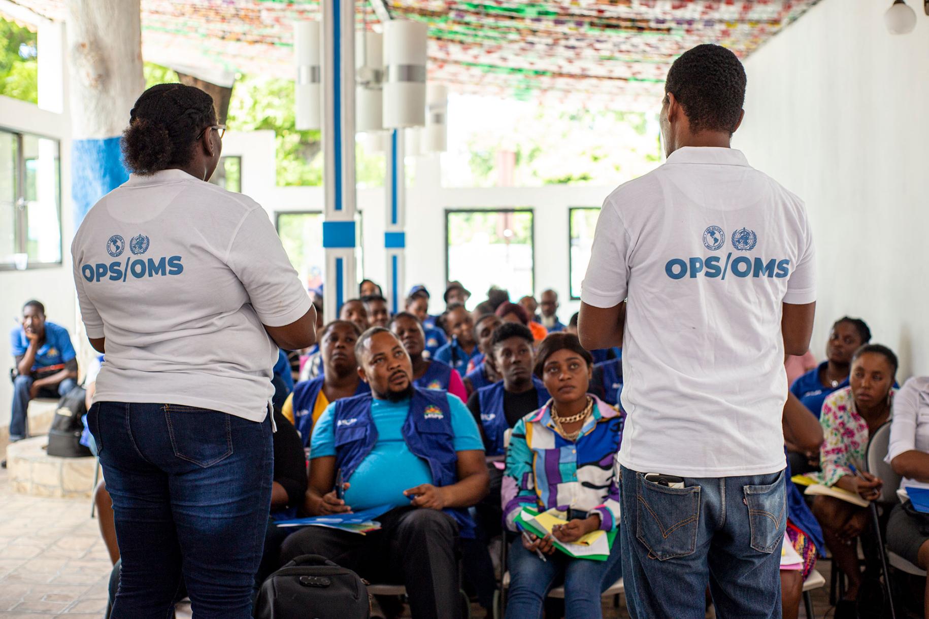 Deux employés de l’OPS/OMS vêtus tous deux d’un jean et d’un t-shirt blanc s’adresse à des agents communautaires de santé dans une salle de formation, en Haïti.