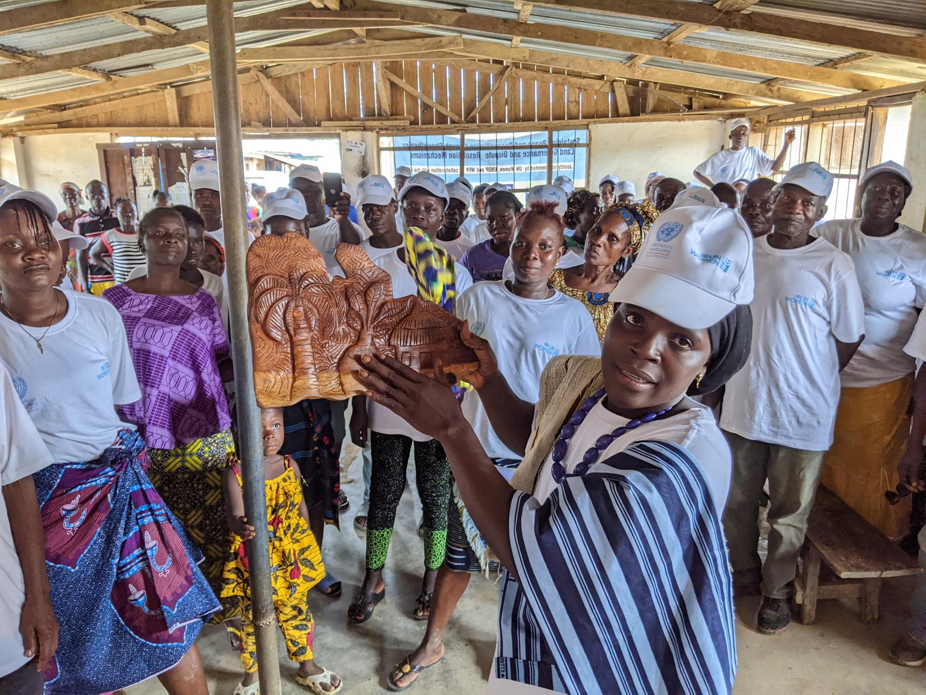 En Liberia, un grupo de mujeres y hombres están reunidos en una estancia y miran hacia la cámara mientras una de las mujeres, en primer plano, muestra una escultura hecha a mano.