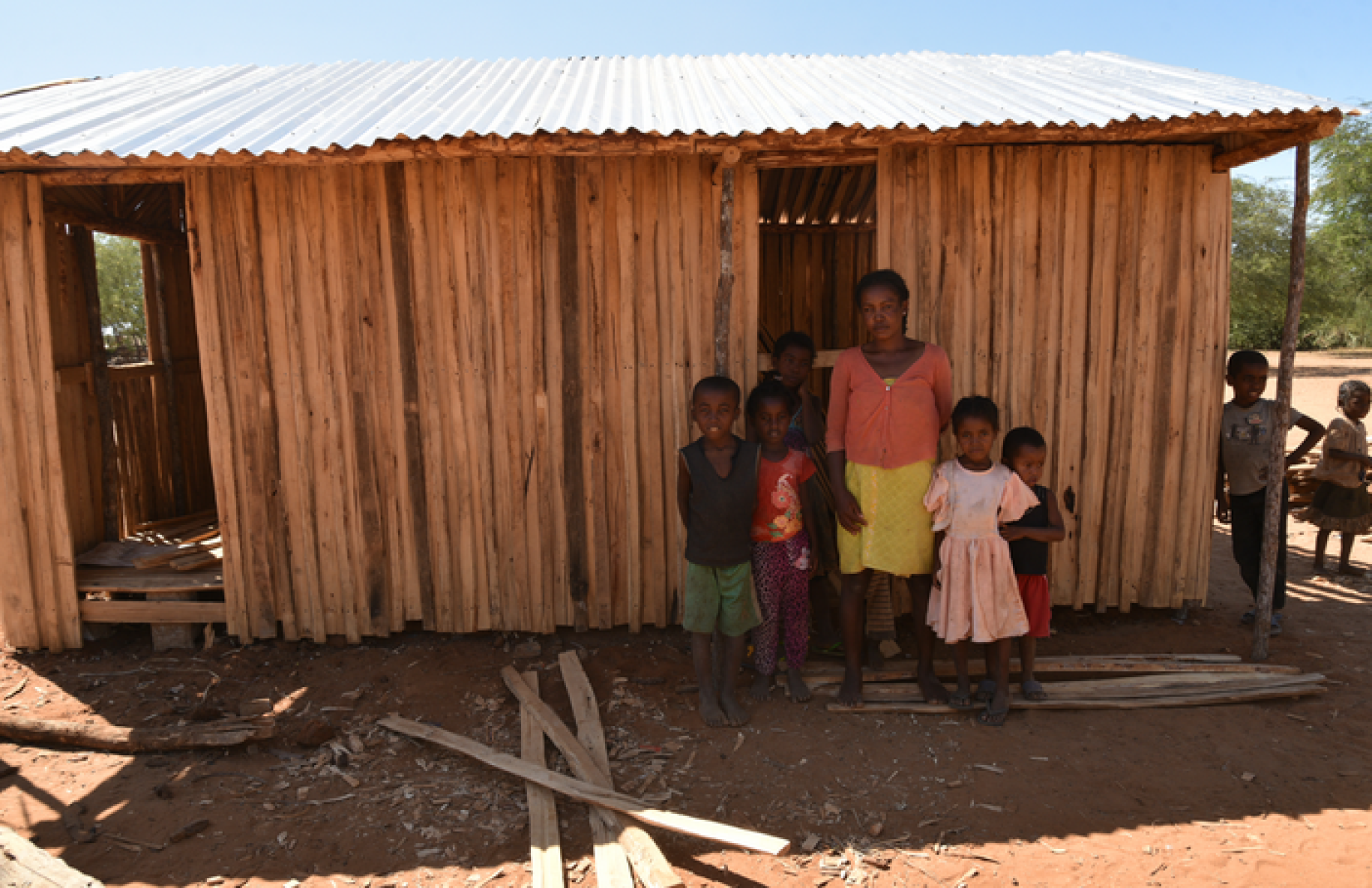 A Malagasy woman stands before the camera, surrounded by young children, in front of a humble wooden house in the village of Amhamahavel, Madagascar.