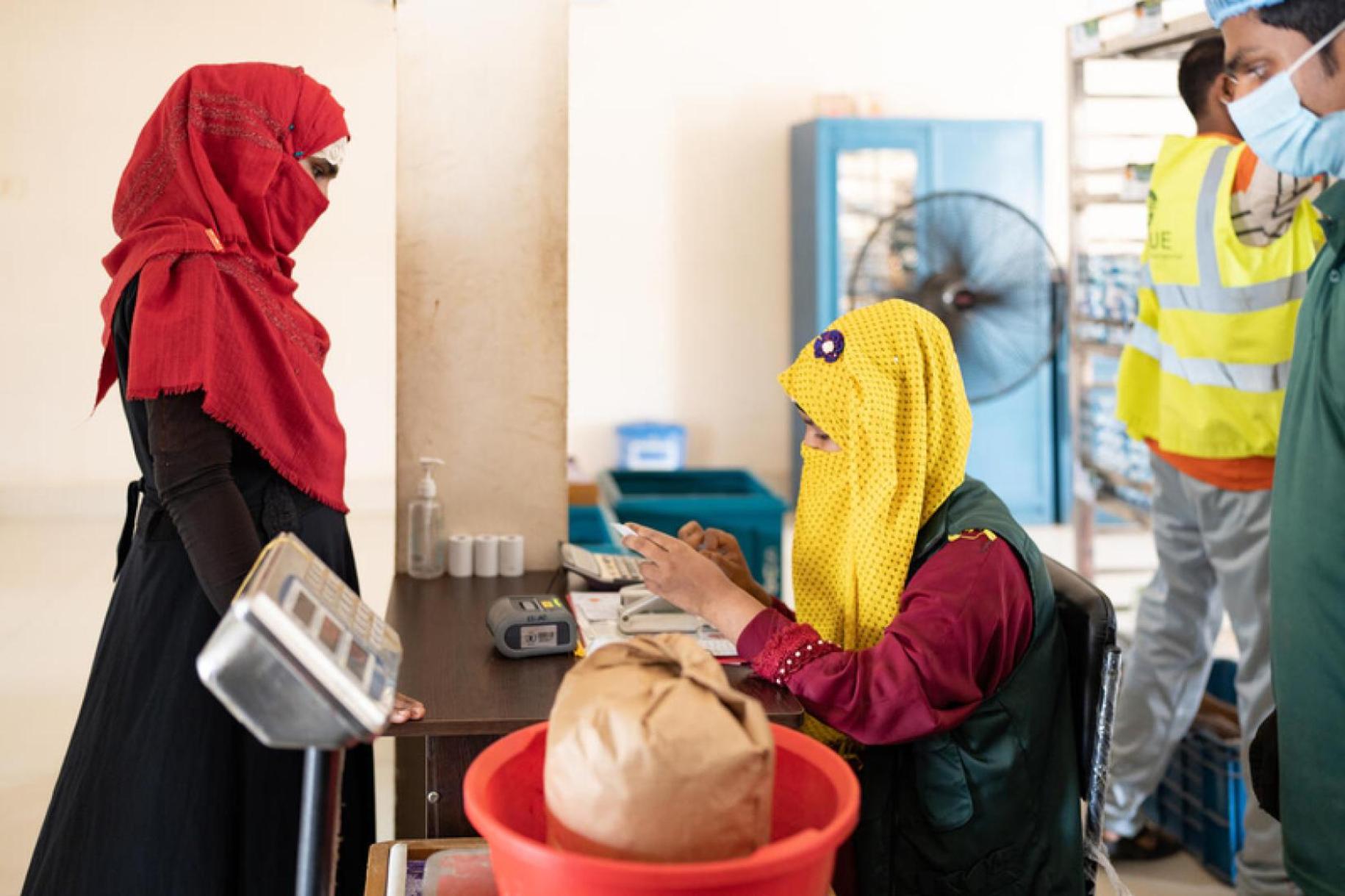 Woman in a a red veil and a black dress stands at a food distribution counter while a woman in a yellow veil and black dress weighs a bag on a weighing scale