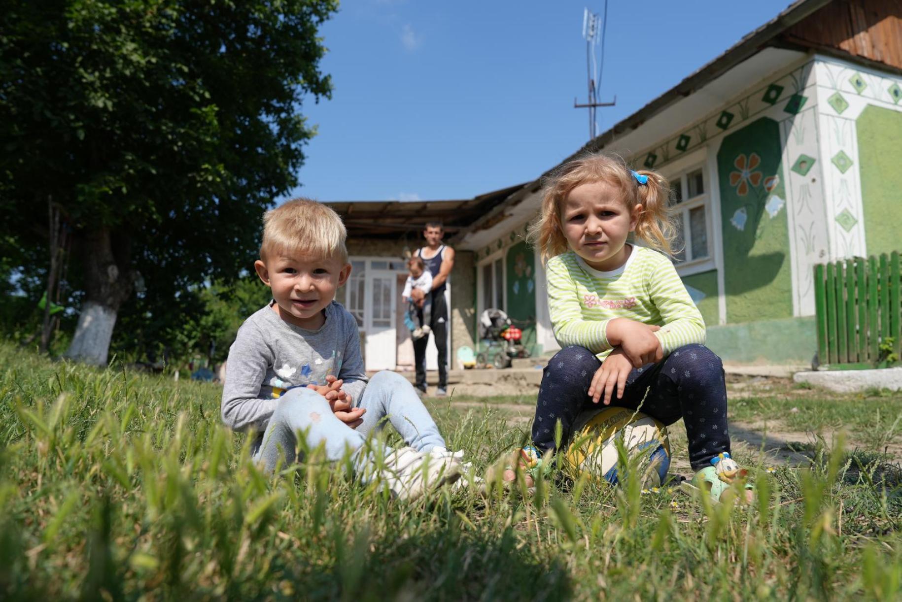 Two children sit on grass in front of a house and smile.