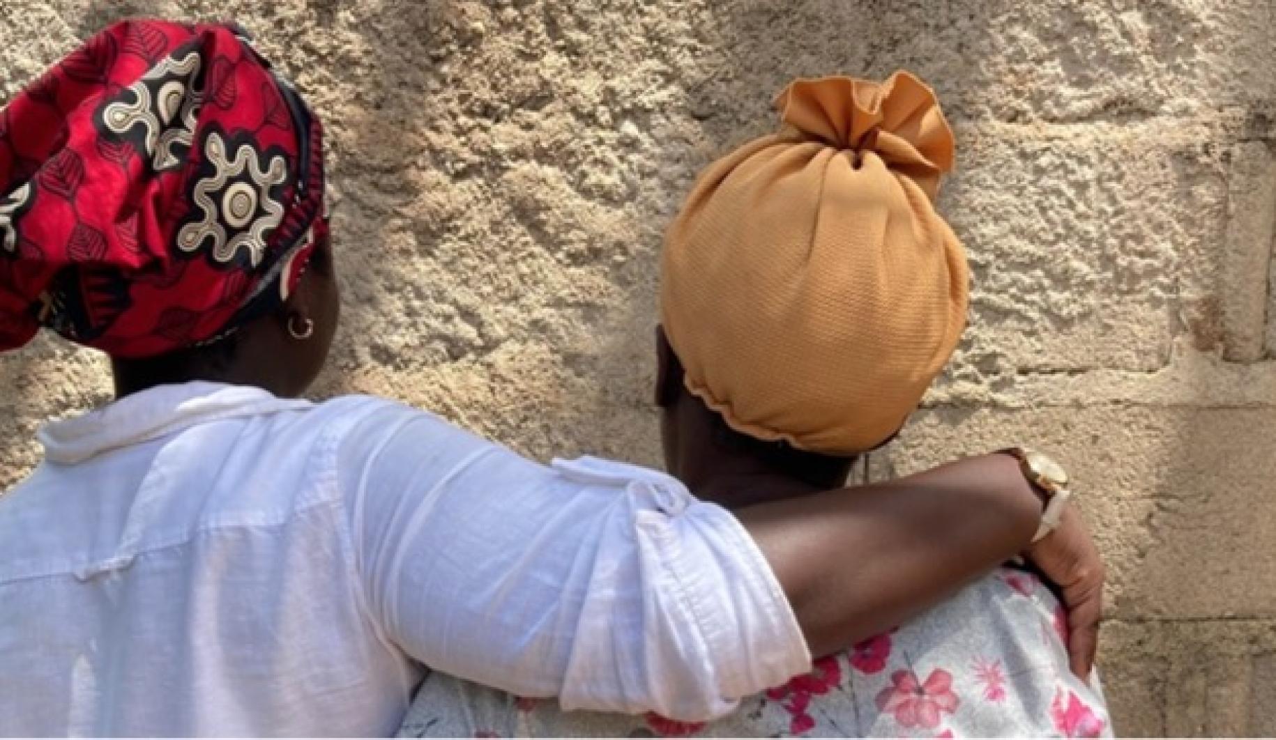 two women in headscarves stand with their backs to the camera, one with her arm over the other 