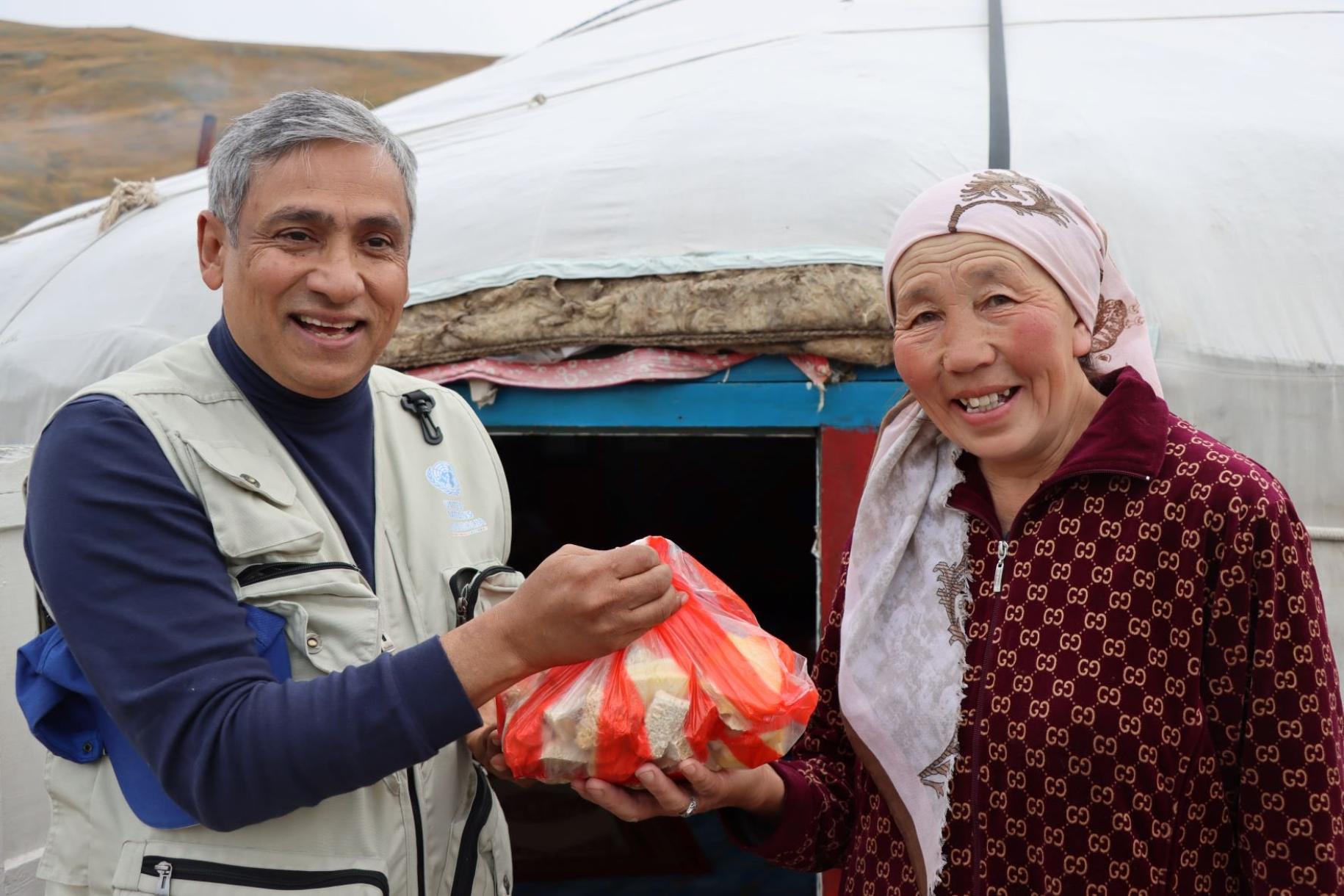 A man in a blue shirt and grey vest hands over an orange coloured bag to a woman in a red dress and headscarf in front of a Yurt in Mongolia