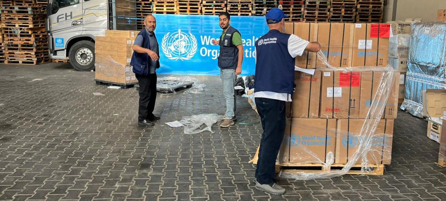 Men unload boxes of supplies and boxes, and stand in front of a World Health Organization sign.