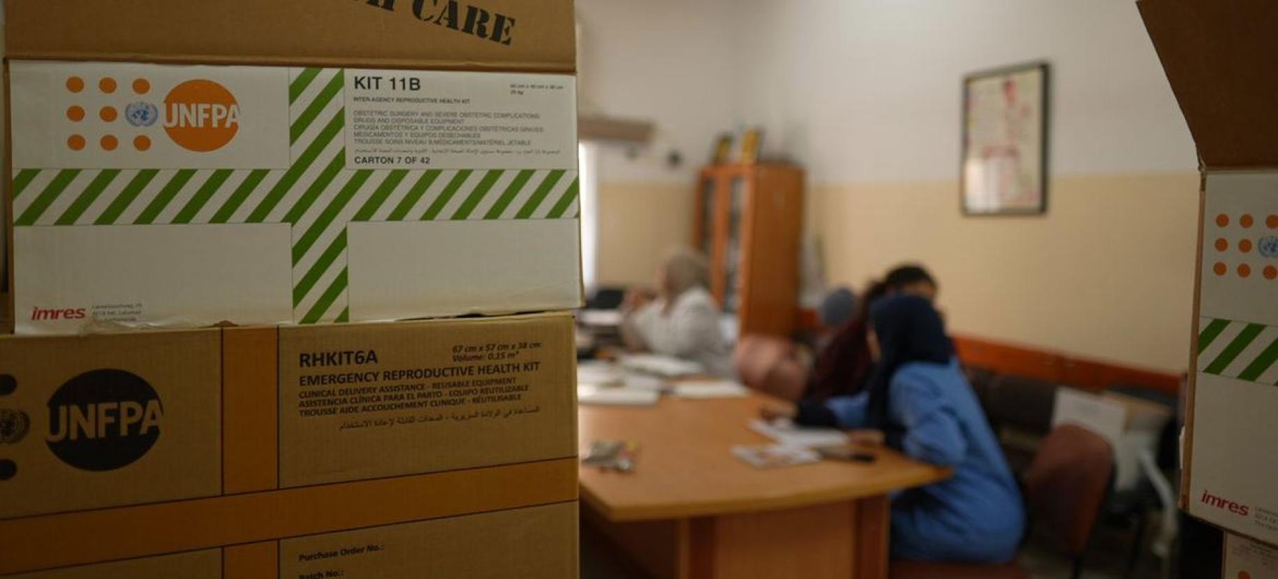UNFPA kits and boxes, with women, not in focus, seated and talking in the background. 