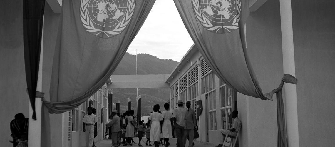 Se muestra a varias personas caminando juntas, alejándose de una entrada, la cual está coronada por dos banderas de las Naciones Unidas.
