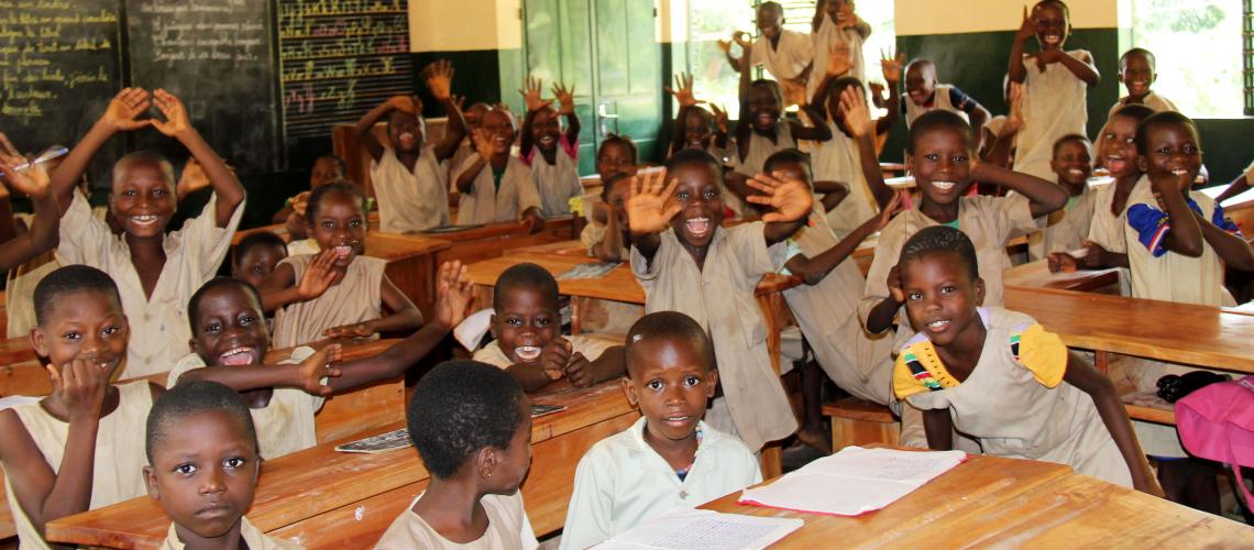 A group of school children smile and wave at the camera inside a classroom.