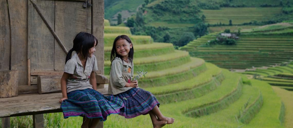 iet Nam banner showing a beautiful green countryside in the background, with two small smiling girls sitting on the porch of a home smiling at each other in the foreground.