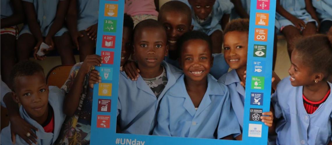 Niños y niñas posan para la cámara. Algunos aparecen sonriendo detrás de un gran marco hecho con cartulina que muestra los logotipos de los ODS.