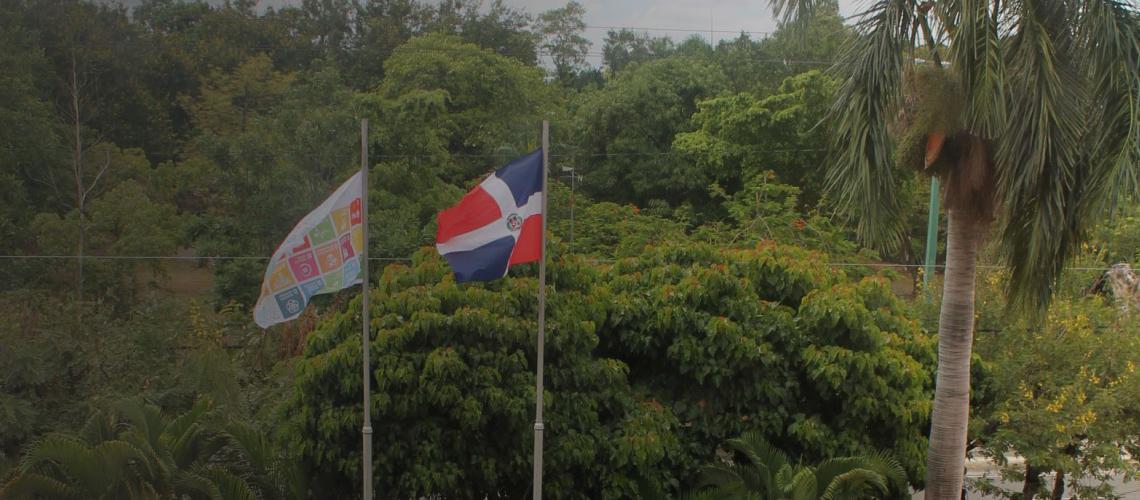 Una bandera de los ODS y la bandera de la República Dominicana ondean en lo alto de una exuberante campiña.