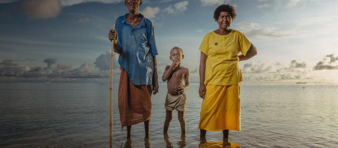 An older man with a cane, young boy and woman stand in shallow water under an illustrious sky above them.