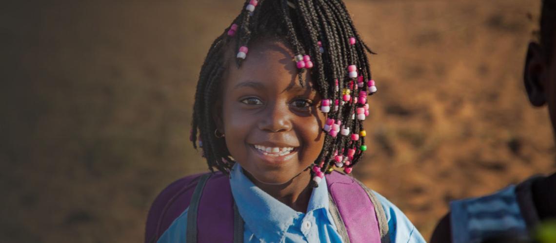 A little girl wearing a backpack smiles happily at the camera. 