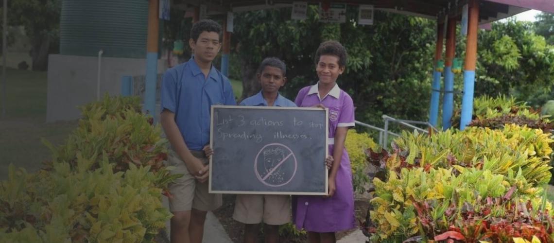 Three young students of the Natawa Primary School hold up a chalkboard outside their school