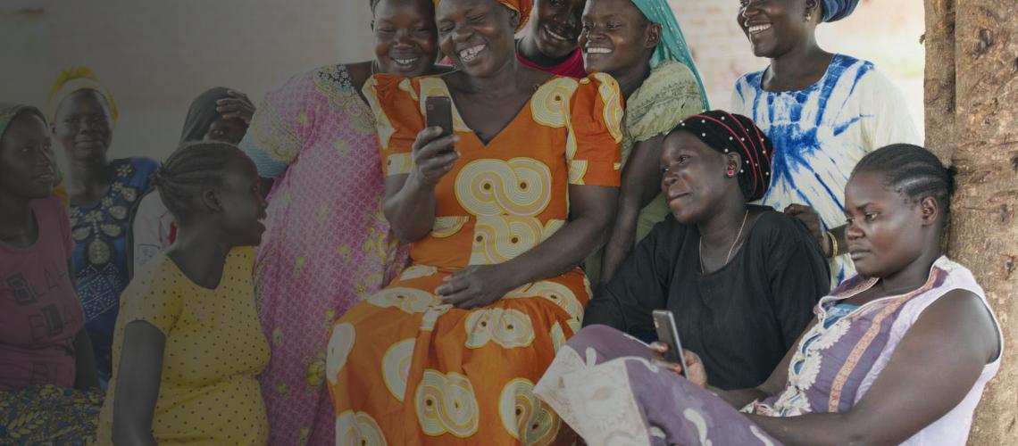 Women happily smile and look at a mobile phone as a group as they surround one woman who holds it.