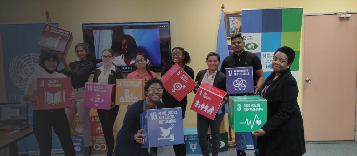 Jóvenes en una sala se sitúan frente a una videollamada y sonríen a la cámara mientras sostienen bloques decorados con los logotipos de los ODS.