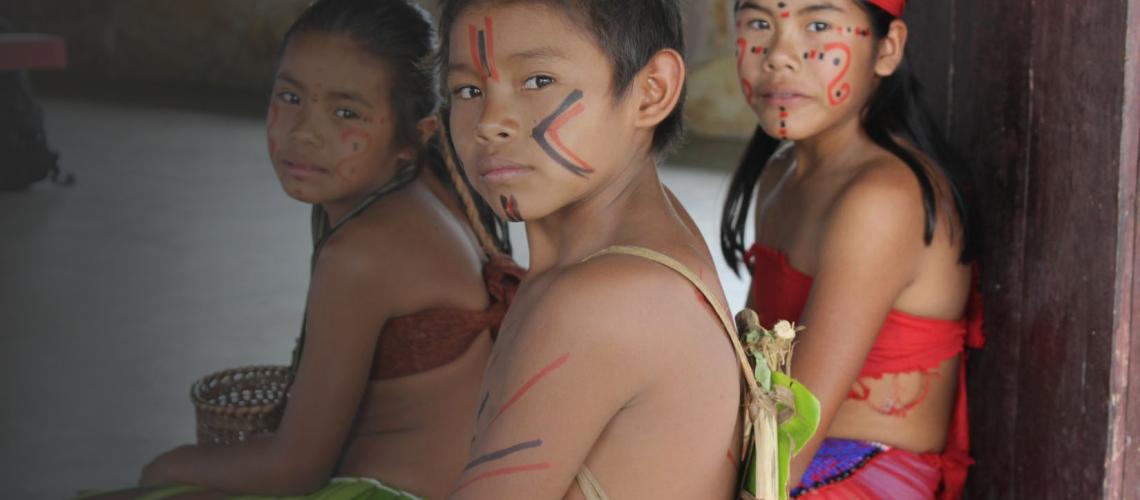 Niños y niñas indígenas, que llevan el rostro pintado y usan trajes tradicionales, se sientan en el suelo mientras miran a la cámara.