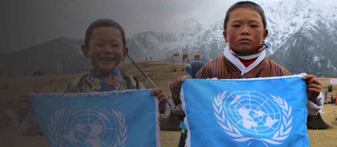 Dos niños, uno sonriente y otro serio, sostienen la bandera azul de las Naciones Unidas con una montaña nevada de fondo.