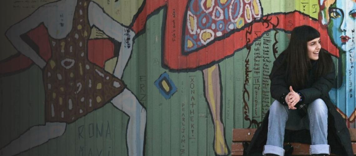 Девушка с длинными темными волосами сидит на фоне стены, расписанной граффити. 