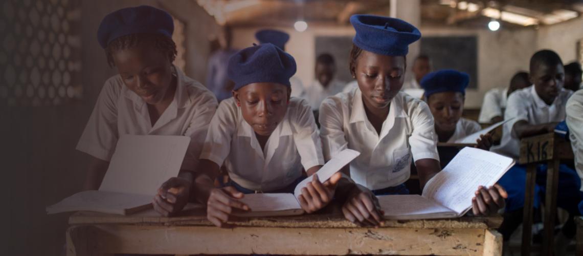 Three young adolescent girls sit together at a table in the front row of their classroom as they studiously read their notebooks.