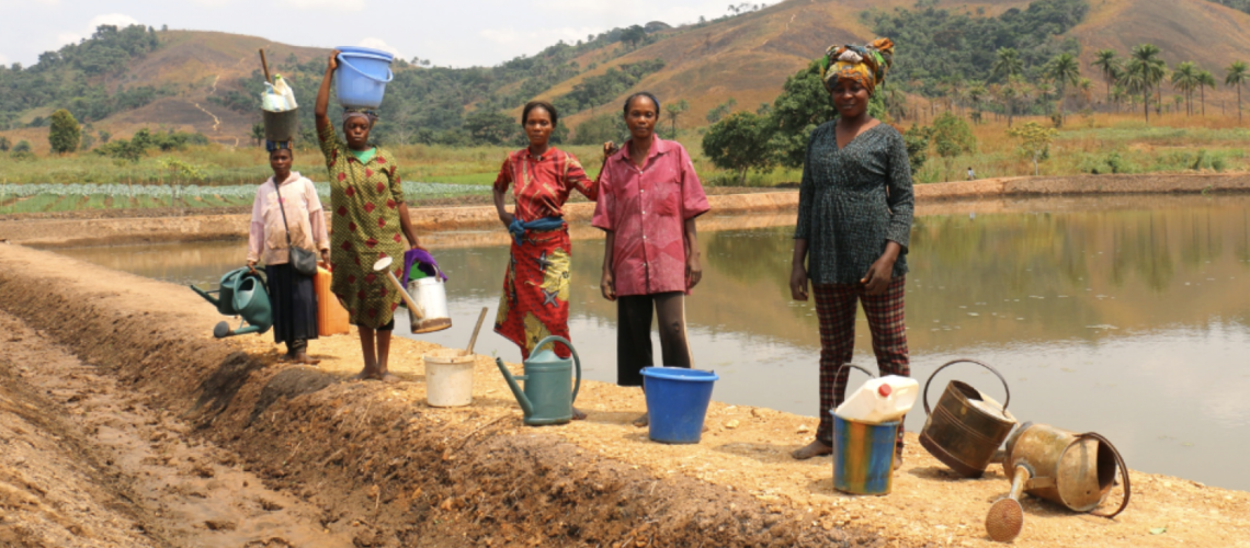 Five women stand near a body of water with buckets both on their heads and on the ground.