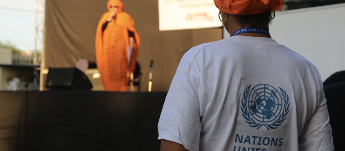 Una imagen de una mujer con un vestido naranja hablando por un micrófono mientras otra mujer, con una camiseta de la ONU en Mauritania, observa. 