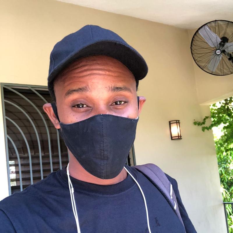 A man in a black face mask, black baseball cap, and black shirt looks directly at the camera. 