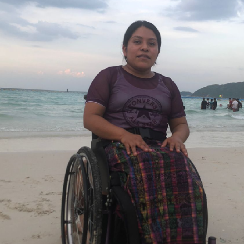 La imagen muestra a Floridalma Bocel Raxtún sentada en una silla de ruedas en una playa de Guatemala